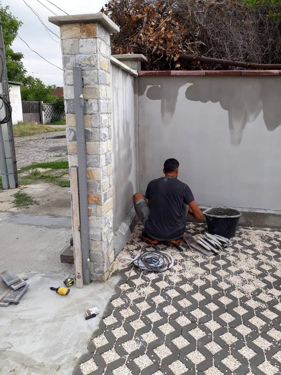 Worker cladding a gate pillar with cut stone beside pebble-filled grid paving