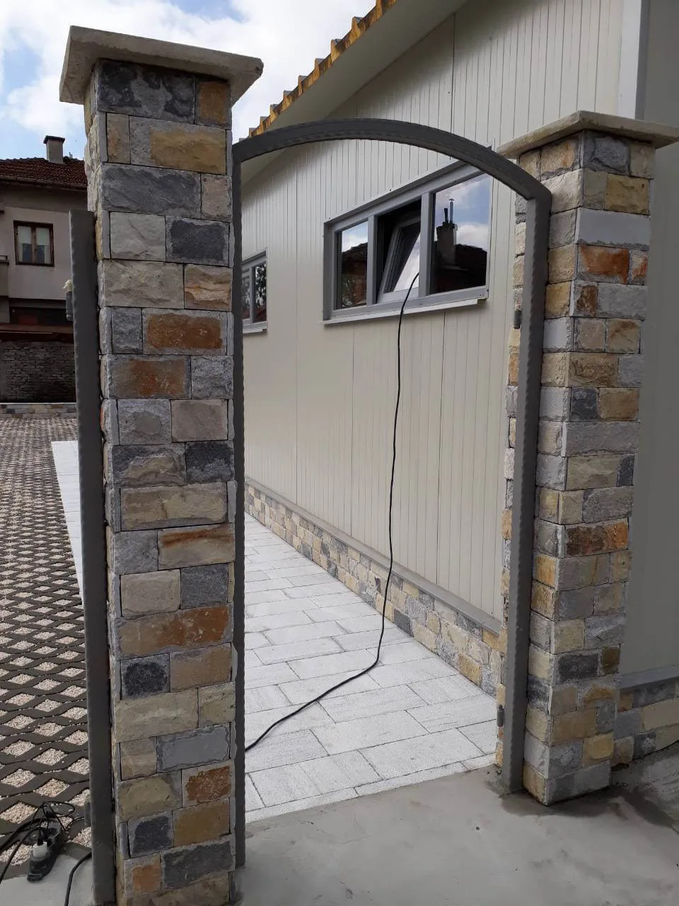 Stone-clad gate pillars with arched metal frame and a paved yard behind