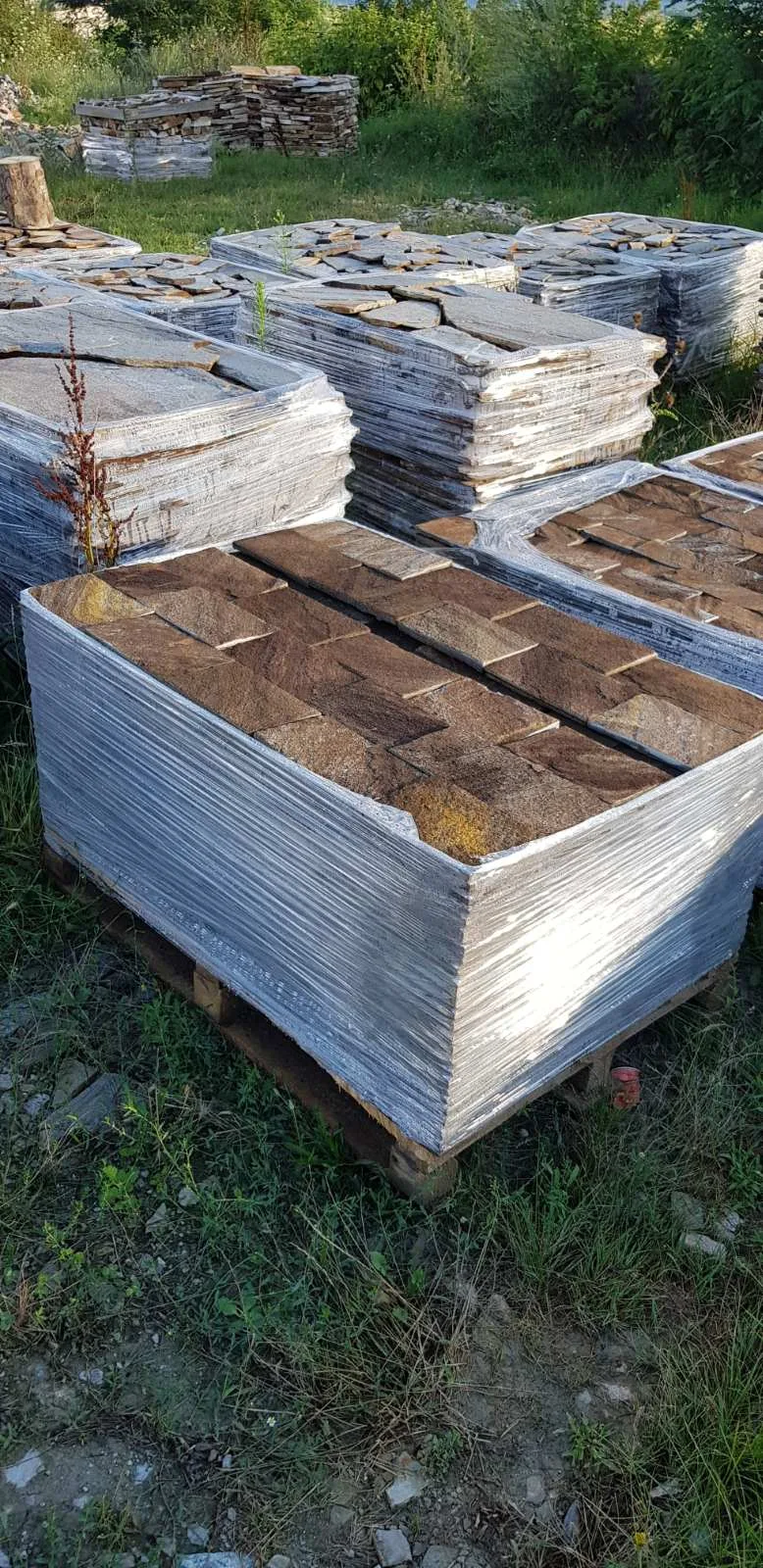 Foil-wrapped pallets of brown rock-faced gneiss cladding tiles in a field yard